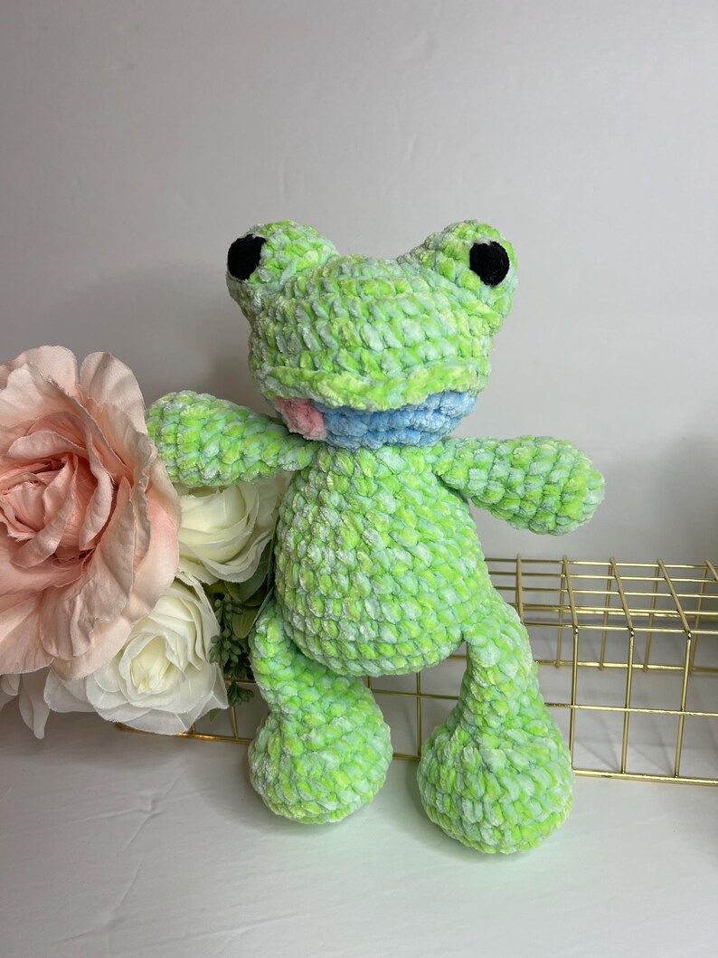 Puede incluir: Una rana de peluche hecha a mano en tonos verdes, azules y rosas. La rana tiene grandes ojos negros y una expresi&oacute;n sonriente. El material suave y texturizado le da a la rana una apariencia tierna. Es un art&iacute;culo artesanal caprichoso.