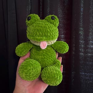 Puede incluir: Un peluche de rana verde hecho a mano con ganchillo, con ojos de seguridad negros y una lengua rosa. La rana tiene un aspecto texturizado y difuso y est&aacute; sentada con las piernas extendidas. El peluche se sostiene en una mano.