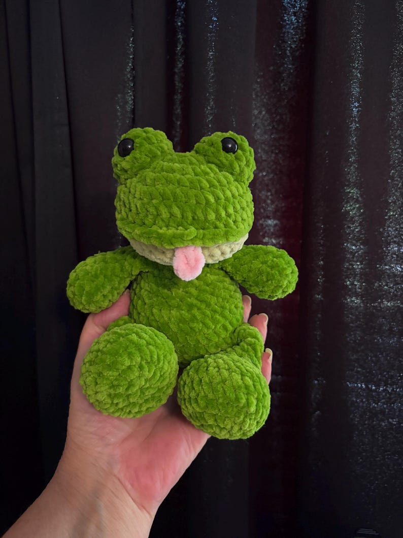 Puede incluir: Un peluche de rana verde hecho a mano con ganchillo, con ojos de seguridad negros y una lengua rosa. La rana tiene un aspecto texturizado y difuso y est&aacute; sentada con las piernas extendidas. El peluche se sostiene en una mano.