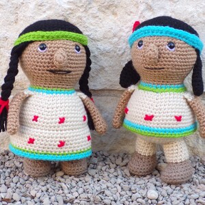Puede incluir: Dos muñecas hechas a mano con ganchillo, con trenzas negras, que visten vestidos color crema con detalles de estrellas rojas y ribetes turquesa y verdes. Una muñeca tiene una diadema verde, la otra una diadema azul. Las muñecas están sobre una superficie de piedra.
