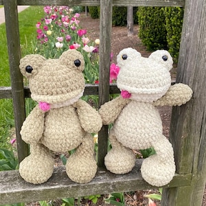 Puede incluir: Dos peluches de rana hechos a mano con ganchillo. Uno es verde claro, el otro blanco roto. Ambos tienen ojos negros y detalles rosas alrededor del cuello. Est&aacute;n sobre una valla de madera con un jard&iacute;n al fondo.