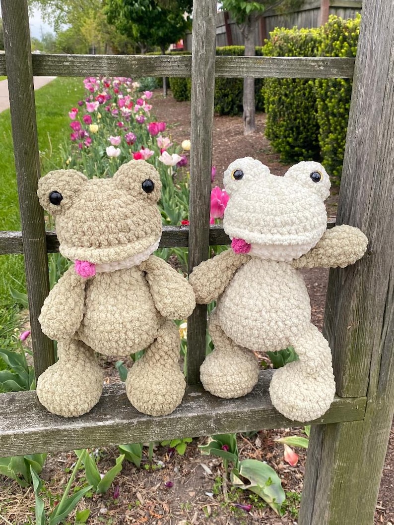 Puede incluir: Dos peluches de rana hechos a mano con ganchillo. Uno es verde claro, el otro blanco roto. Ambos tienen ojos negros y detalles rosas alrededor del cuello. Est&aacute;n sobre una valla de madera con un jard&iacute;n al fondo.