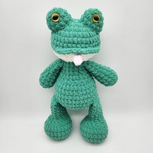 Puede incluir: Un peluche de rana verde hecho a mano con ganchillo, con grandes ojos amarillos y una lengua rosa. La rana tiene un cuello blanco y est&aacute; de pie. El peluche est&aacute; hecho de un hilo suave y texturizado.
