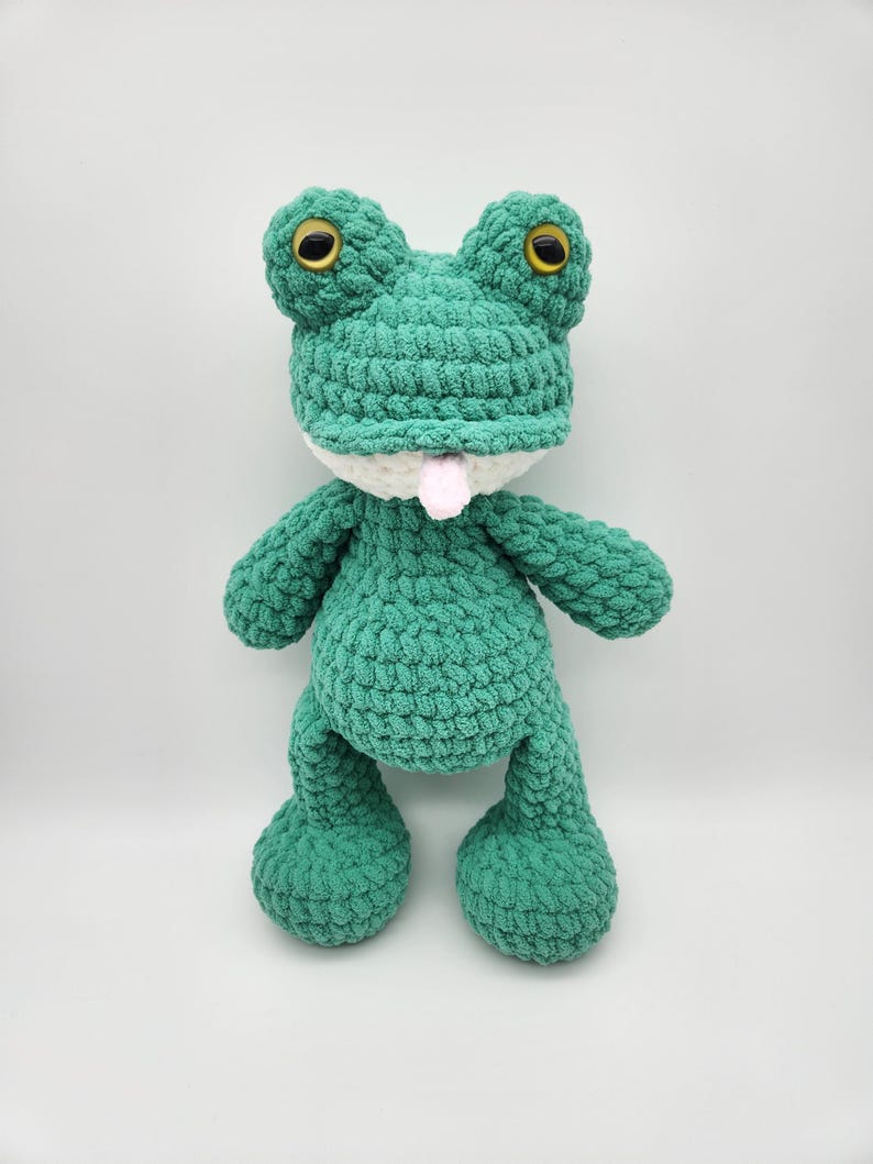 Puede incluir: Un peluche de rana verde hecho a mano con ganchillo, con grandes ojos amarillos y una lengua rosa. La rana tiene un cuello blanco y est&aacute; de pie. El peluche est&aacute; hecho de un hilo suave y texturizado.