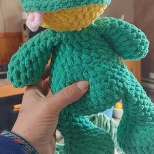 Puede incluir: Un juguete de rana de peluche hecho a mano en tonos verdes y amarillos. La rana tiene ojos negros grandes, una lengua rosa y est&aacute; hecha de un hilo suave y texturizado. Una persona sostiene la rana.