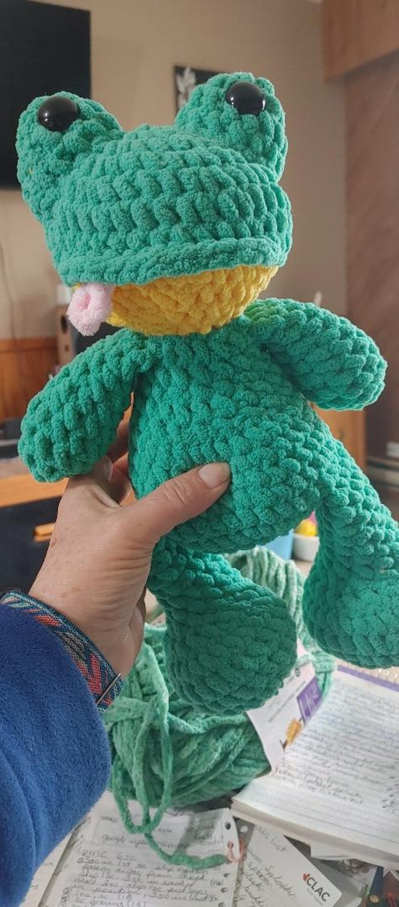 Puede incluir: Un juguete de rana de peluche hecho a mano en tonos verdes y amarillos. La rana tiene ojos negros grandes, una lengua rosa y est&aacute; hecha de un hilo suave y texturizado. Una persona sostiene la rana.