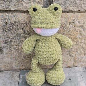 Puede incluir: Un peluche de rana de ganchillo verde claro hecho a mano, con ojos negros y una lengua rosa. La rana tiene una boca blanca y est&aacute; hecha de un hilo suave y texturizado. La rana est&aacute; sobre una superficie de madera.