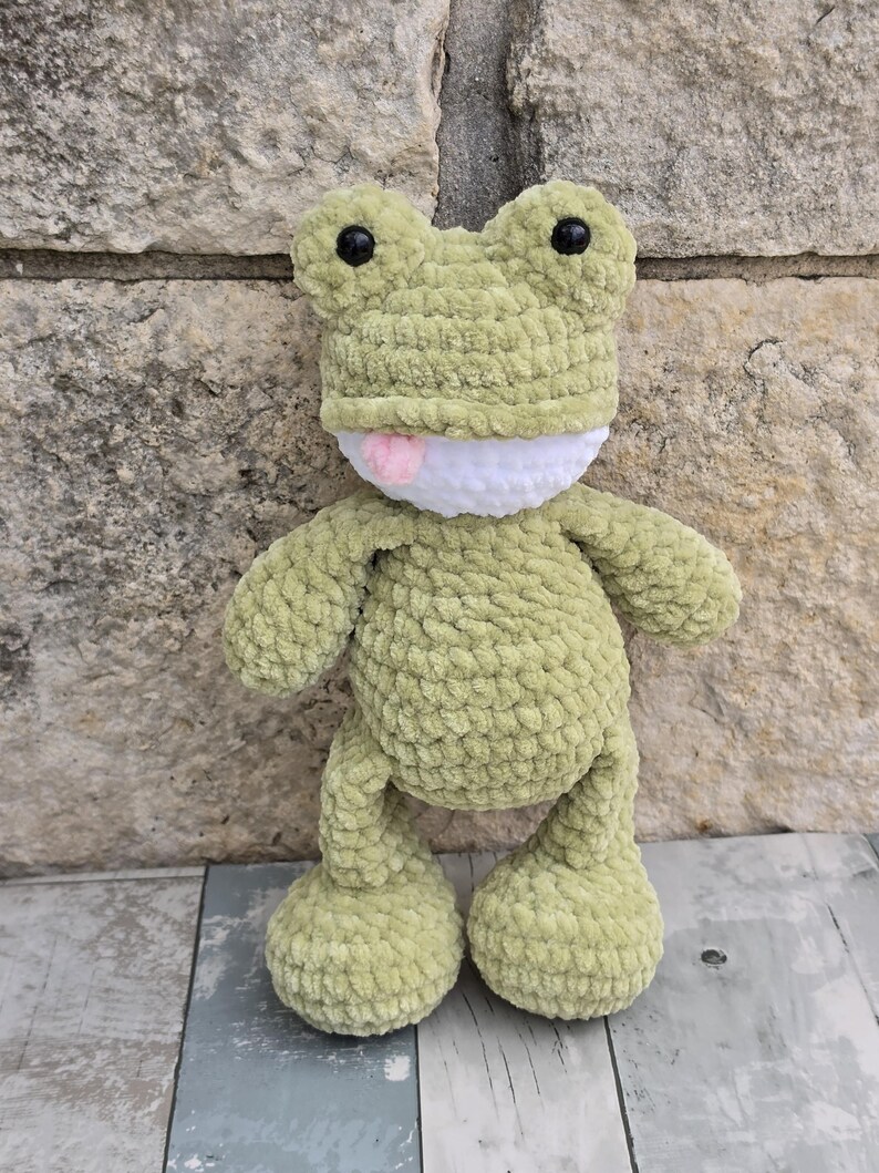 Puede incluir: Un peluche de rana de ganchillo verde claro hecho a mano, con ojos negros y una lengua rosa. La rana tiene una boca blanca y est&aacute; hecha de un hilo suave y texturizado. La rana est&aacute; sobre una superficie de madera.