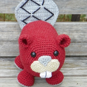 May include: A handmade crochet beaver toy in shades of red, gray, and cream. The beaver has a red body, gray tail, and cream-colored teeth and snout. Black safety eyes and small ears complete the design. A charming, soft toy.