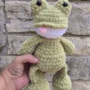 Puede incluir: Un peluche de rana de ganchillo verde claro hecho a mano, con ojos negros y una lengua rosa. La rana tiene una barriga blanca y se sostiene contra una pared de piedra. El peluche es suave y texturizado.