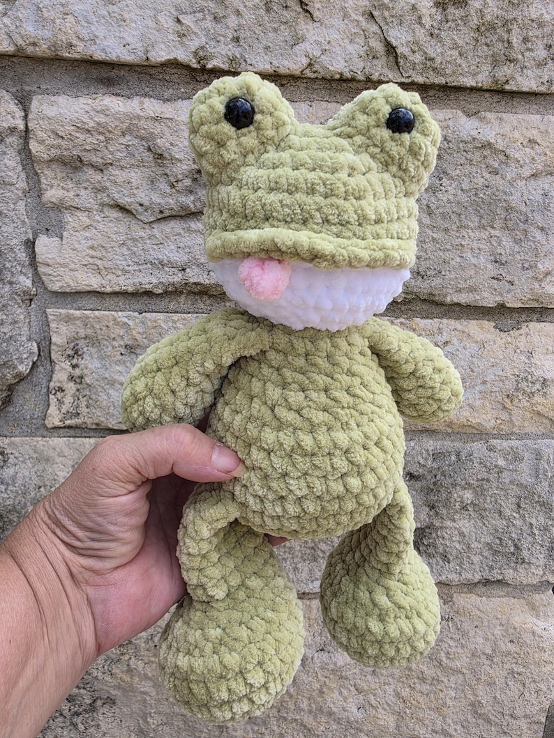 Puede incluir: Un peluche de rana de ganchillo verde claro hecho a mano, con ojos negros y una lengua rosa. La rana tiene una barriga blanca y se sostiene contra una pared de piedra. El peluche es suave y texturizado.