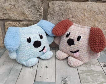 May include: Two handmade crochet dog-shaped pillows. One is light blue with blue ears, the other is light pink with brown ears. Both have black eyes, noses, and smiling mouths with pink tongues. They are on a light-coloured wooden surface.