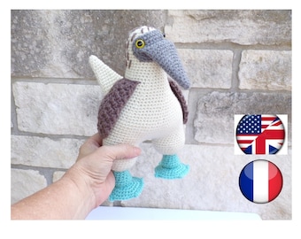 May include: A crocheted blue-footed booby bird toy with a white body, brown wings, and blue feet. The toy is being held in a person's hand.