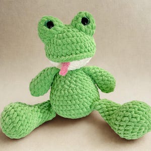 Puede incluir: Un peluche de rana verde hecho a mano con ganchillo, con ojos negros grandes y una lengua rosa. La rana tiene un cuello blanco y est&aacute; sentada con las piernas extendidas. El peluche est&aacute; hecho de un hilo suave y texturizado.