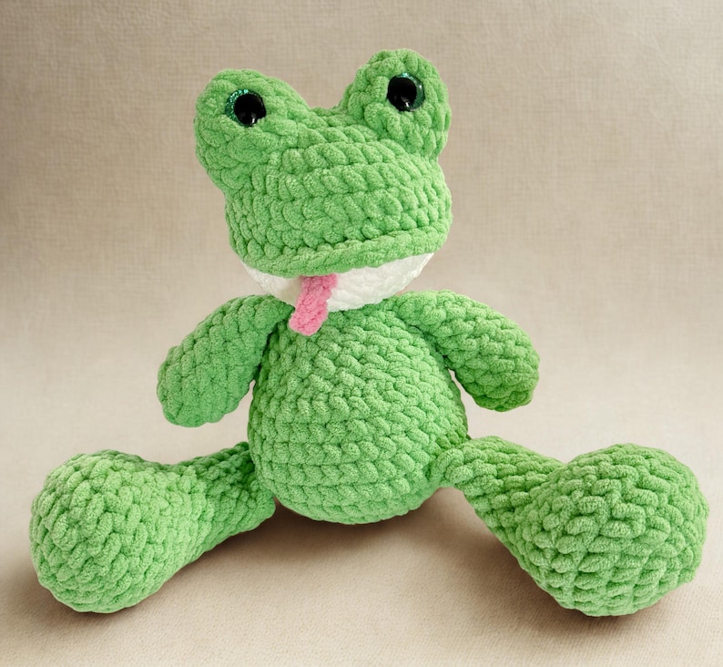 Puede incluir: Un peluche de rana verde hecho a mano con ganchillo, con ojos negros grandes y una lengua rosa. La rana tiene un cuello blanco y est&aacute; sentada con las piernas extendidas. El peluche est&aacute; hecho de un hilo suave y texturizado.