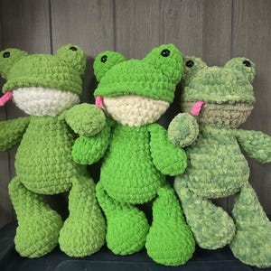 Puede incluir: Tres peluches de rana hechos a mano con ganchillo en varios tonos de verde. Cada rana tiene una cara blanca, ojos negros y una lengua rosa. Los peluches son suaves y texturizados, con una apariencia caprichosa y amigable.