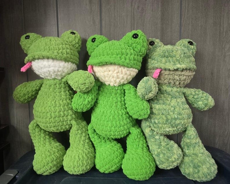 Puede incluir: Tres peluches de rana hechos a mano con ganchillo en varios tonos de verde. Cada rana tiene una cara blanca, ojos negros y una lengua rosa. Los peluches son suaves y texturizados, con una apariencia caprichosa y amigable.