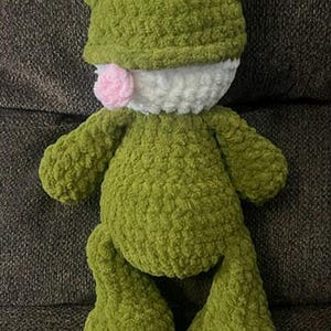 Puede incluir: Un juguete de peluche de rana verde hecho a mano con ojos negros, boca blanca y lengua rosa. La rana tiene un aspecto texturizado y difuso y est&aacute; sentada sobre una superficie marr&oacute;n oscuro.