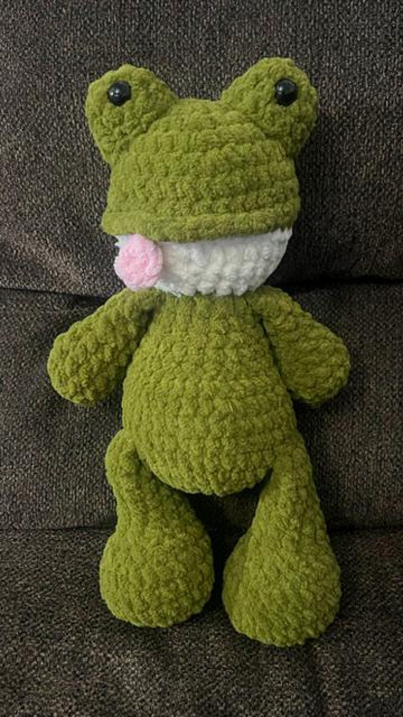 Puede incluir: Un juguete de peluche de rana verde hecho a mano con ojos negros, boca blanca y lengua rosa. La rana tiene un aspecto texturizado y difuso y est&aacute; sentada sobre una superficie marr&oacute;n oscuro.