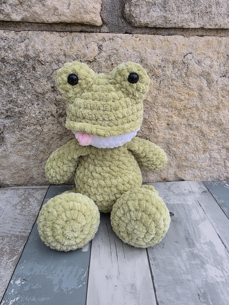 Puede incluir: Un peluche de rana de ganchillo verde claro hecho a mano con ojos negros y una lengua rosa. La rana tiene una boca blanca y est&aacute; sentada sobre una superficie de madera. El peluche es un art&iacute;culo suave y tierno.
