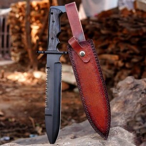 Könnte beinhalten: Ein feststehendes Messer mit mattschwarzer Oberfläche, gezacktem Messerrücken und einem Griff in dunkler Holzoptik. Das Messer wird von einer rotbraunen Lederscheide mit geprägten Mustern und einem einzelnen Metalldruckknopf begleitet.