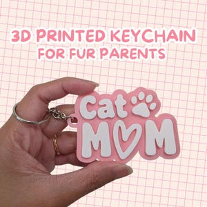 May include: A pink and white 3D printed keychain featuring the text Cat Mom with a heart shape replacing the letter O and a paw print icon. The keychain is attached to a silver metal split ring for keys, showcasing a layered design.