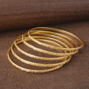 Peut inclure: Un ensemble de cinq bracelets joncs fins en or, chacun présentant un motif en relief délicat et répétitif sur la surface extérieure. Les bracelets sont circulaires et possèdent une finition métallique polie, reflétant la lumière.