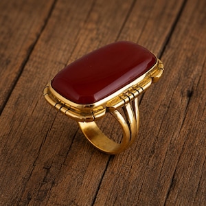Può includere: Un anello color oro con una grande pietra cabochon rettangolare rosso scuro incastonata in una montatura di metallo lucido. La fascia è composta da tre fili paralleli che si uniscono in un gambo solido, sostenendo la liscia pietra rossa.
