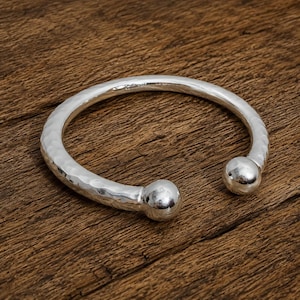 May include: A silver-toned open cuff bracelet featuring a hammered, textured surface along the band and two smooth, polished spherical ball ends. The metal has a reflective finish, and the design is circular with a gap between the two ends.