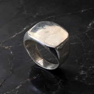 Puede incluir: Un anillo de sello de plata maciza con una cara cuadrada lisa y pulida y una banda gruesa y redondeada. La superficie metálica presenta un brillo reflectante con sutiles variaciones naturales en la textura a lo largo de toda la estructura.