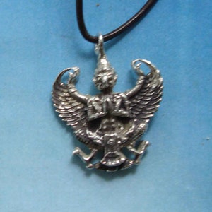 Puede incluir: Colgante plateado con un diseño detallado de Garuda, suspendido de un cordón negro. El Garuda, una criatura mítica con forma de ave, se representa con alas extendidas y detalles intrincados. El colgante probablemente está hecho de metal y se muestra sobre un fondo azul.