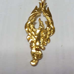 May include: A gold-coloured pendant necklace featuring a detailed design of a mythical creature with wings and a tail. The pendant is attached to a gold-coloured chain.