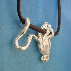 Könnte beinhalten: Ein silberner Affenanhänger, der an einem braunen Lederband hängt. Der Affe ist dargestellt, wie er kopfüber an einem Ast hängt.