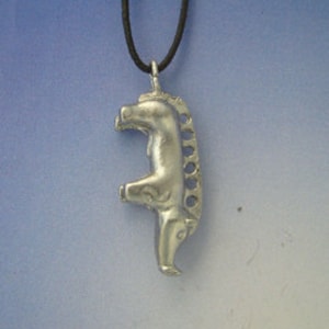 Puede incluir: Collar con colgante de plata con un diseño de caballo estilizado. El caballo está de pie sobre sus patas traseras con la cabeza girada hacia un lado. El colgante está unido a un cordón negro.