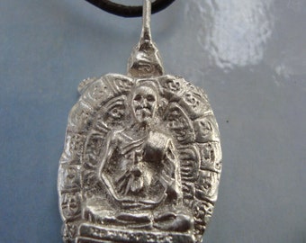 May include: Silver pendant featuring a detailed depiction of a seated Buddha figure surrounded by intricate designs. The pendant is attached to a black leather cord.
