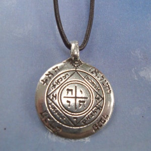Può includere: Una collana con un ciondolo d'argento su un cordoncino di cuoio nero. Il ciondolo ha un design circolare con testo ebraico e una croce al centro.