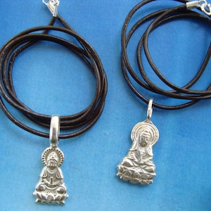 Puede incluir: Dos collares de plata con cordones de cuero marrón y colgantes con una figura de Buda sentado.