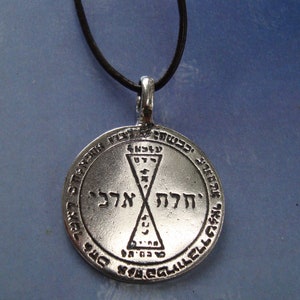 May include: A silver pendant necklace with a black cord. The pendant features a circular design with Hebrew text and a geometric symbol.