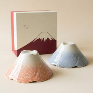 May include: Two ceramic bowls shaped like Mount Fuji, one with a pink gradient and one with a blue gradient, both featuring a speckled finish. Behind them sits a square gift box with a graphic illustration of a mountain and the text Fuji Japan.