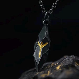 Könnte beinhalten: Ein dunkler, facettierter, geometrischer Anhänger an einer passenden dunklen Metallkette. Der Anhänger hat eine texturierte, mattschwarze Oberfläche mit einem markanten, erhabenen goldfarbenen Symbol, das einem stilisierten Y oder einer Rune ähnelt.