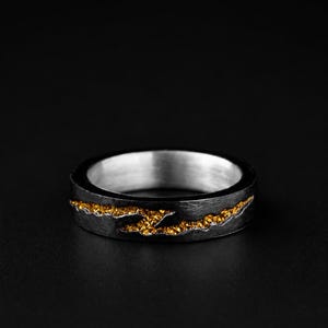 May include: A band ring featuring a dark, textured exterior surface with a horizontal, jagged inlay of shimmering gold-colored granular material. The interior of the ring is smooth and polished with a bright silver-toned metallic finish.