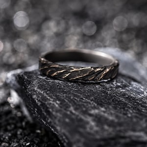 Könnte beinhalten: Ein Ring aus dunklem Metall mit einer texturierten, rauen Oberfläche, tiefen Rillen und erhabenen Graten. Das Band zeigt ein kontrastreiches Finish mit dunkelgrauen Tönen und subtilen metallischen Kupferakzenten auf der unebenen Außenseite.