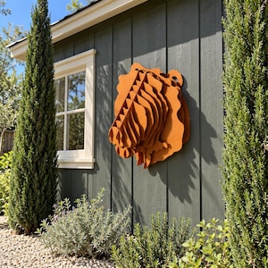 Può includere: Una scultura da parete tridimensionale a forma di testa di leone, realizzata con strati di materiale color ruggine. Il design presenta una serie di pannelli verticali sovrapposti che creano un profilo geometrico stilizzato di un leone.