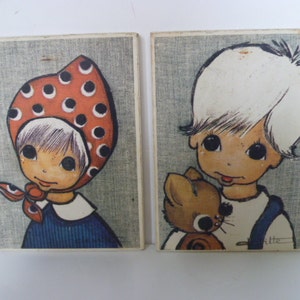 May include: Two framed illustrations of children. The girl is wearing a red and white polka dot hat and a blue dress. The boy is wearing a white shirt and blue overalls and holding a brown stuffed animal.