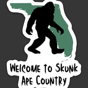 May include: A die-cut sticker featuring a black silhouette of a Bigfoot-like creature walking in front of a muted green map of Florida. Below the map, the text reads "WELCOME TO SKUNK APE COUNTRY" in a jagged, black, hand-drawn style font.