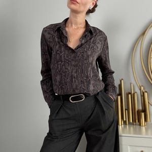 May include: A long-sleeved button-down shirt featuring a dark grey and black snakeskin pattern, paired with charcoal grey trousers and a black belt with a large, oval-shaped silver buckle. The shirt has a classic collar and a buttoned front placket.