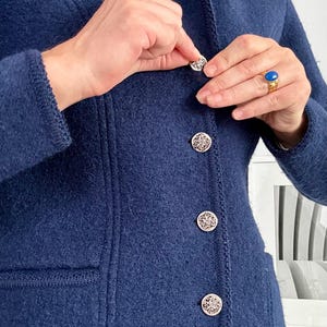 May include: A dark blue boiled wool jacket featuring a high collar and a front button closure. The jacket has four round, silver-toned metal buttons with an intricate embossed pattern, long sleeves, and two front pockets with ribbed trim edges.