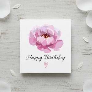 Può includere: Biglietto di auguri quadrato con un'illustrazione di una peonia rosa. Le parole "Happy Birthday" sono scritte in corsivo nero sotto il fiore, con un piccolo cuore rosa sotto il testo. Il biglietto è su una superficie di legno bianco.