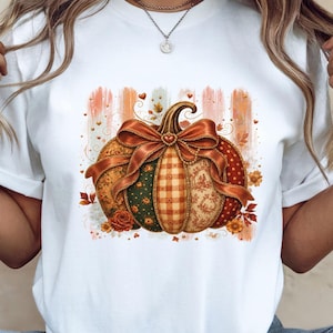 May include: A white short-sleeve t-shirt featuring a central graphic of a pumpkin decorated with a large orange bow. The pumpkin has segments with varied patterns including plaid, floral, and polka dots in shades of orange, green, and cream.