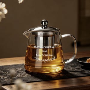 May include: A clear glass teapot with a stainless steel lid and infuser, filled with amber tea. The glass is engraved with the text TOP Grandad Ever, Love, Amy & William, accompanied by a small leaf branch graphic design element.